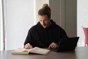 University student reading an open textbook beside a laptop while studying at a desk