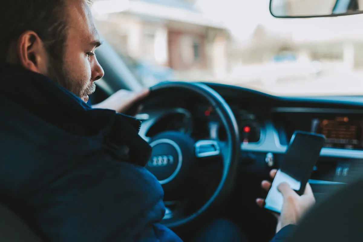 A driver in a dark jacket sitting in the driver seat of a car holding a smartphone with a blank dark screen