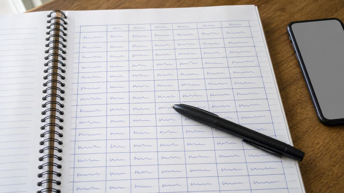 An open spiral-bound kilometre logbook with hand-written entries beside a pen and a smartphone on a wooden kitchen table