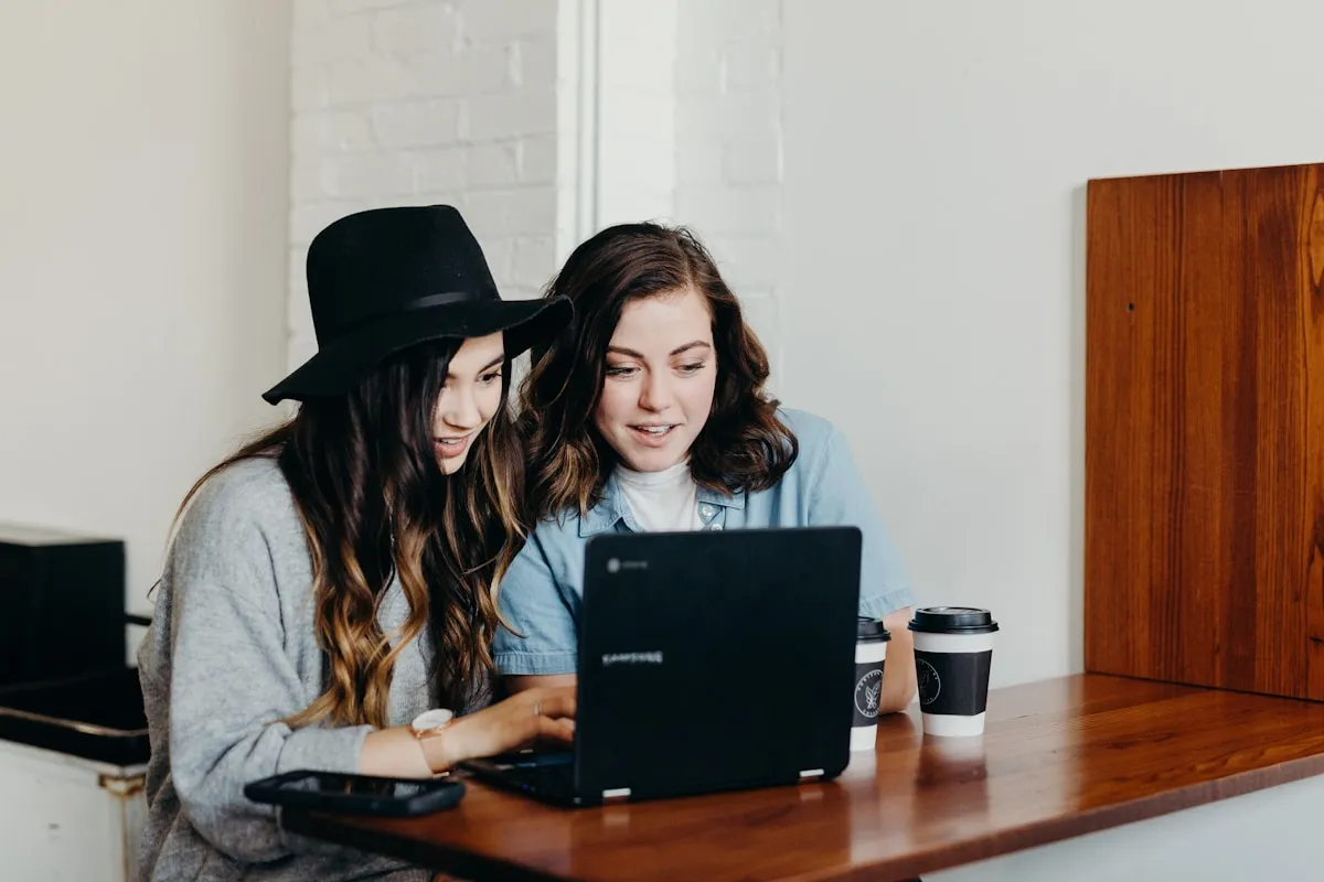 Two young women review a university computer science shortlist together at a cafe table over an open laptop