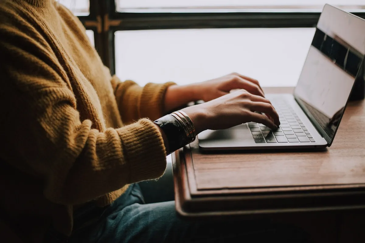 Close up of a student typing on a laptop at a wooden desk beside a sunlit window in a yellow sweater