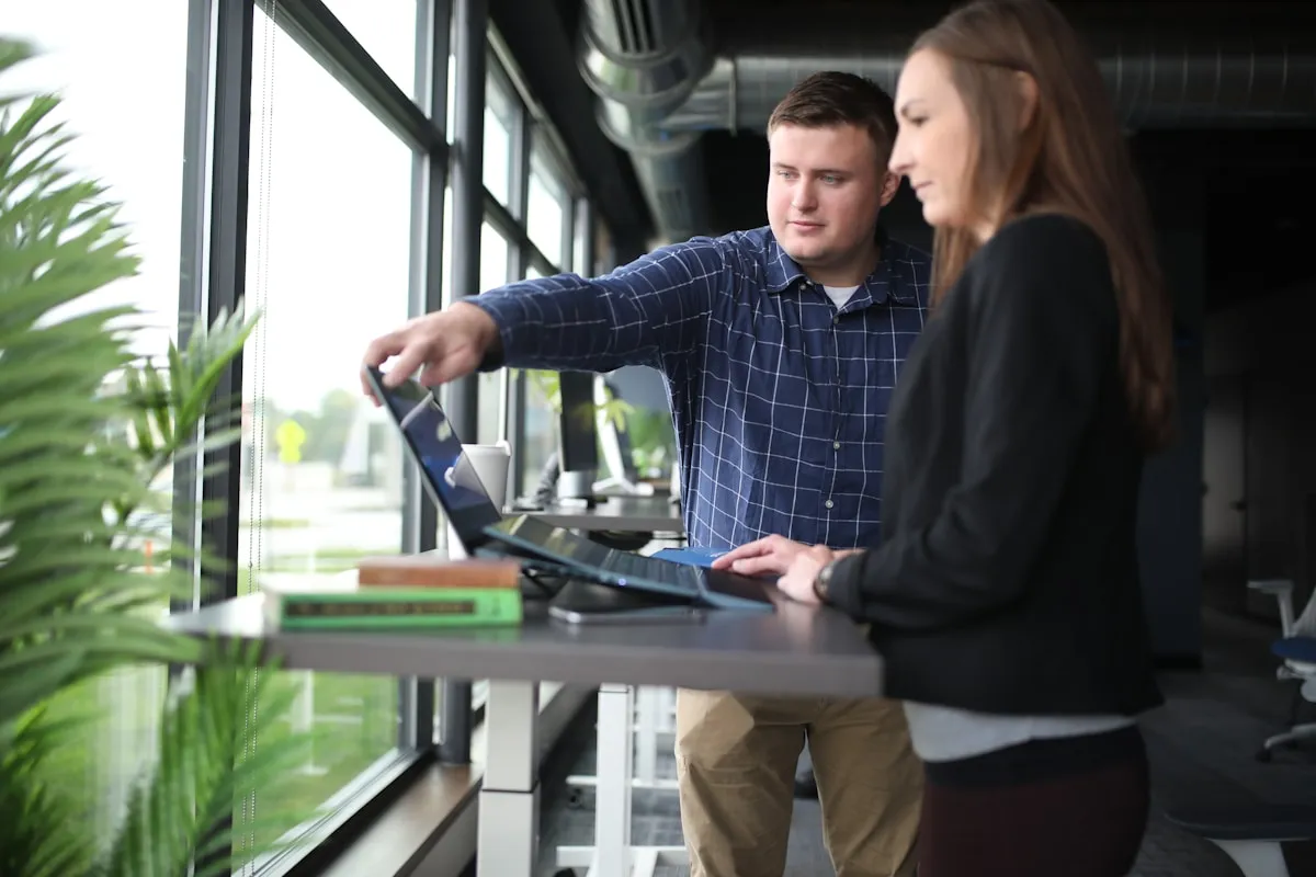 A young man points at a laptop screen while a young woman beside him takes notes at a standing desk in a bright office